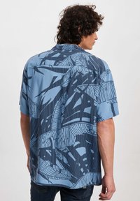 Camicia a maniche corte di colore azzurro chiaro con motivi geometrici e botanici blu scuro. Tessuto morbido con una vestibilità comoda e orlo diritto.