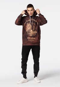 Brun hoodie med en illustration av Crater Lake National Park i Oregon, med detaljer av träd och lodge. Bärs med svarta byxor och vita skor.