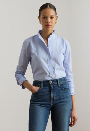 Γυναίκα με τα χέρια στις τσέπες, φορώντας φανελάκι με ελαφρύ γαλάζιο βολάν στον γιακά και σκούρο μπλε τζιν ψηλόμεσο, μπροστά σε απλό φόντο.