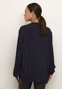 Blouse marine à manches longues avec des rayures verticales argentées, un col froncé et une coupe ample. Elle présente des manches larges et un ourlet légèrement arrondi.