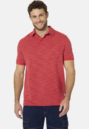 Man with curly hair smiling, wearing a red short-sleeve polo shirt and dark blue cargo pants, standing against a white background.