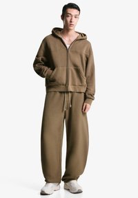 Brun zip-up hættetrøje med frontlommer og matchende brede sweatsbukser. Stoffet er blødt og har en afslappet pasform. Hvide sneakers fuldender looket.