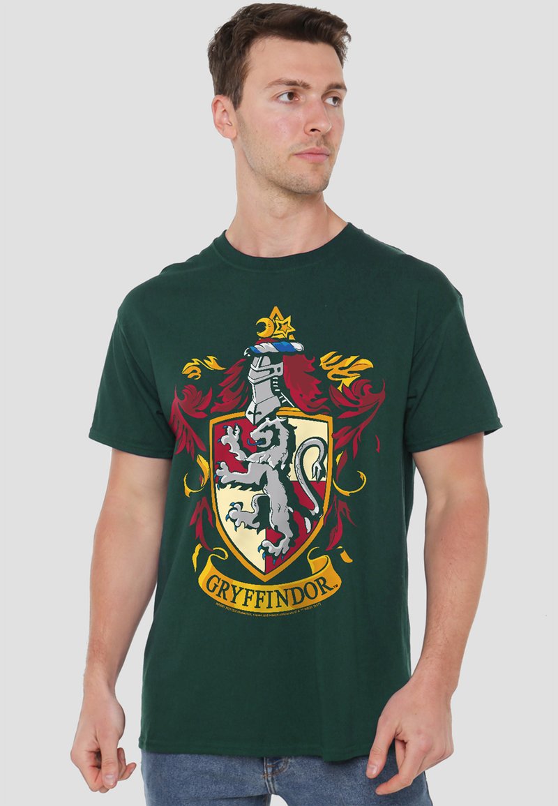 Young man wearing a dark green Gryffindor crest t-shirt, standing and looking to his left against a plain light background.