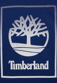 Modrá tkanina s bílým logem Timberland, zobrazující stylizovaný strom uvnitř kruhového obrysu, s názvem značky pod ním.