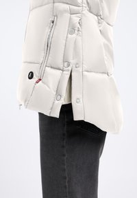 Jasnoszara kurtka puchowa z pikowanym wzorem, z zatrzaskami i zapięciem na zamek. Posiada boczne szczeliny i kontrastową czerwoną metkę.