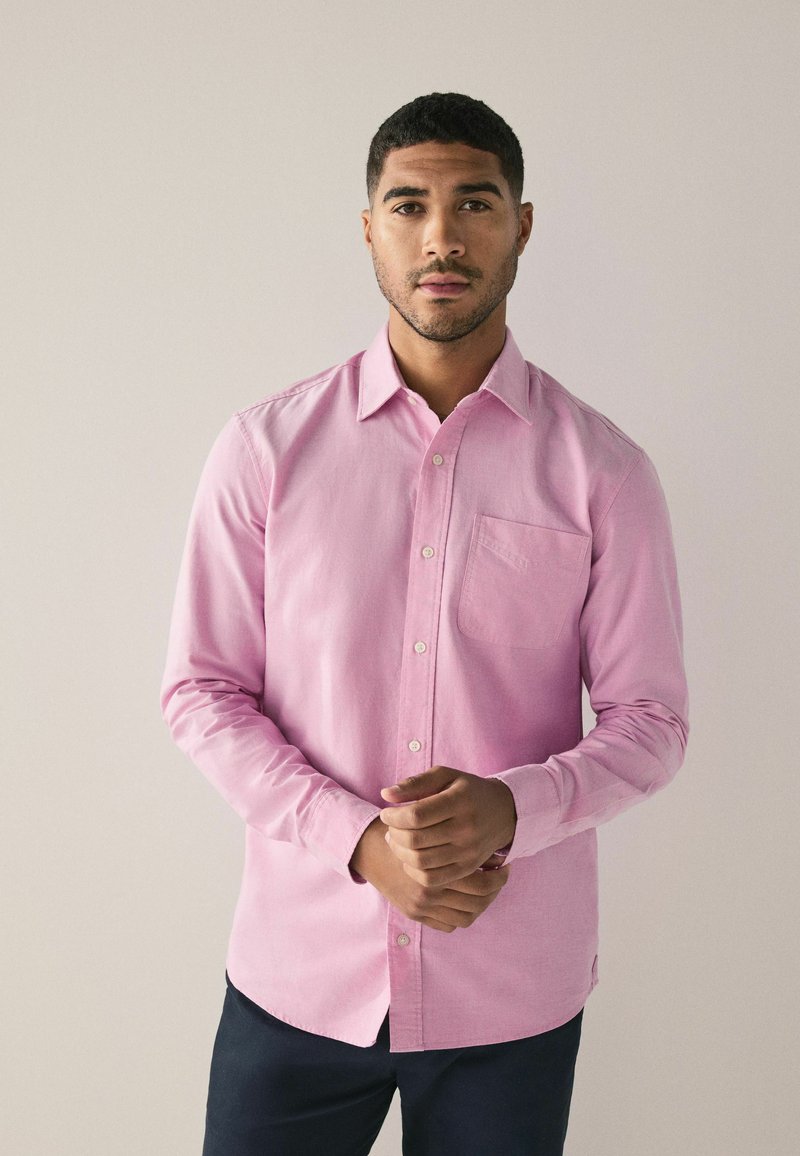 Helles rosa Hemd mit Knöpfen aus einem weichen Stoff, mit einer Brusttasche, langen Ärmeln und einem spitzen Kragen, kombiniert mit dunklen Hosen.