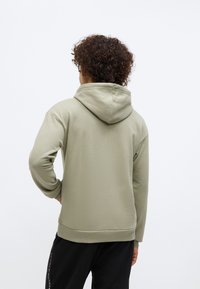 Osoba s kučeravými vlasmi, oblečená v svetlozelenom mikine s kapucňou a čiernych nohaviciach od Tommy Hilfiger, otočená chrbtom s jednou rukou na páse.