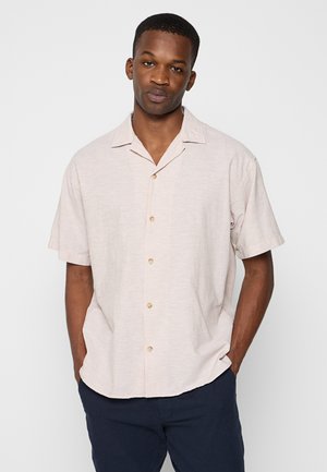 Giovane uomo con capelli corti che indossa una camicia beige chiaro a maniche corte con bottoni e pantaloni scuri, in piedi con le mani in tasca davanti a uno sfondo bianco.