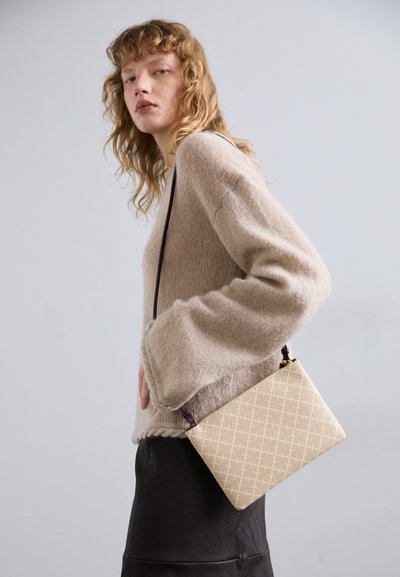 Sac à bandoulière beige avec une texture a motifs, forme rectangulaire et bandoulière marron. Porté avec un pull beige confortable et une jupe noire.