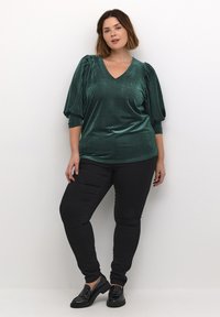 Smaragdgrøn ribbet top med V-hals og puffede ærmer, parret med sorte stramme jeans og glossy sorte sko, sat mod en neutral baggrund.