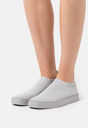 Voeten die lichtgrijze gebreide slip-on sneakers dragen met witte rubberen zolen tegen een witte achtergrond, tonen de onderbenen en enkels.