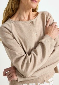 Beige svetr s kruhovým výstřihem z měkké látky s knoflíkovou légou, žebrovanými manžetami a bočními rozparky. Design je volného střihu a hladké textury.