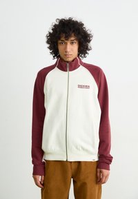 White and burgundy zip-up jacket with a stand collar, ribbed cuffs, and a logo on the chest. Textured fabric with a relaxed fit.