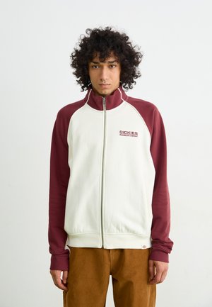 White and burgundy zip-up jacket with a stand collar, ribbed cuffs, and a logo on the chest. Textured fabric with a relaxed fit.