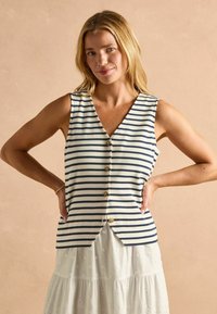 Bezrukávový pásikavý top v krémovej a námorníckej modrej farbe, s výstrihom do V, zapínaním na gombík a bočnými rozparkami, skombinovaný s bielou vrstvenou sukňou.
