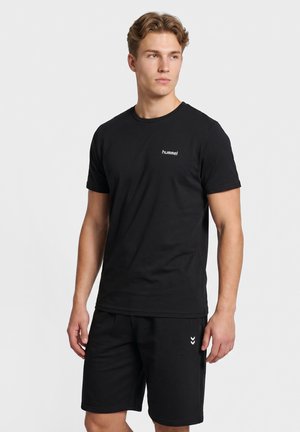 Junger Mann trägt schwarzes hummel T-Shirt und schwarze Shorts, steht vor einfachem hellem Hintergrund und schaut zur Seite.