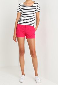 Pruhované tričko s krátkým rukávem v bílé a námořnické modři, v kombinaci s jasně růžovými šortkami na míru. Bílé tenisky dotvářejí celý outfit.