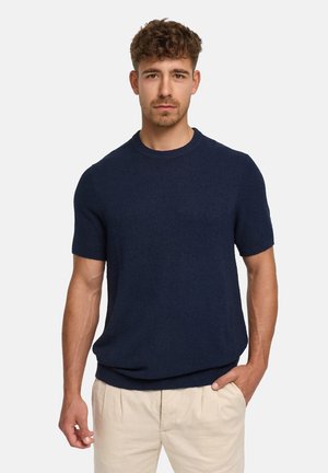 Mann mit lockigem Haar, der einen dunkelblauen Kurzarmpullover und beige Hosen trägt, steht mit einer Hand in der Tasche vor einem einfarbigen Hintergrund.