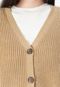 Strikket beige cardigan med V-hals, med et teksturert mønster og to brune knapper foran. Laget over en hvit ribbet topp.