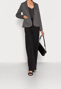Kvinna som bär en grå rutiga blazer, svart linne med spetskanter, breda svarta byxor, svarta flätade sandaler och håller en svart handväska med guldkedja.