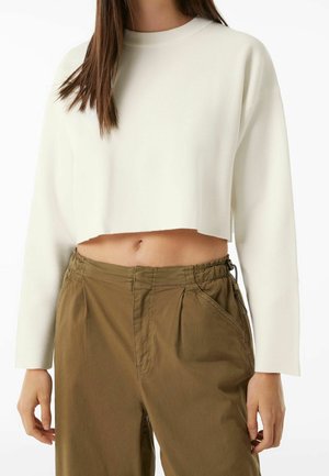 Femme portant un pull court blanc à manches longues et un pantalon ample marron taille haute, debout devant un fond blanc.