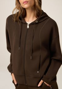 Felpa con zip marrone, dotata di cappuccio, due tasche frontali, hardware argentato e cuciture a contrasto. Tessuto morbido con vestibilità rilassata.