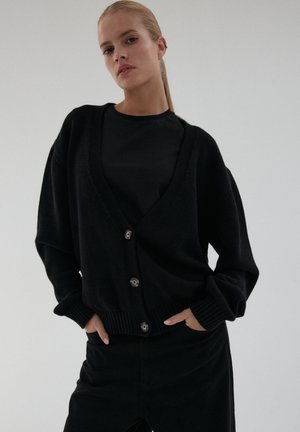 Kobieta z blond włosami w kucyku nosi czarny kardigan zapinany na guziki na czarnej koszulce i czarnych spodniach, pozując z rękami w kieszeniach.