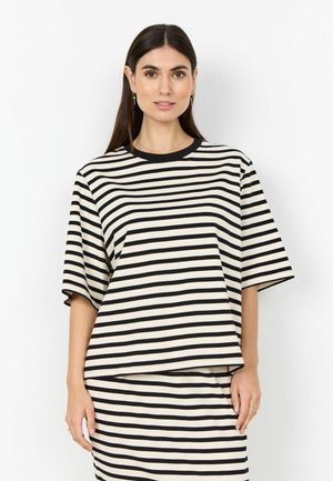Kvinde med langt mørkt hår iført sort og hvidt kortærmet t-shirt med horisontale striber og matchende nederdel, stående foran en hvid baggrund.
