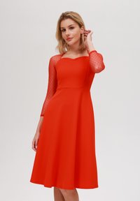Robe rouge avec un corsage ajusté et une jupe évasée, présentant des manches trois-quarts en maille transparente à pois et un col festonné.