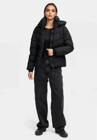 Chaqueta acolchada negra con cuello alto, cierre de cremallera en la parte delantera y capucha con cordón, combinada con unos pantalones oscuros de corte holgado y zapatos negros.