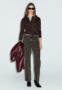 Braun-langärmliges Poloshirt, graues T-Shirt darunter, olivgrüne gerade Jeans, bordeauxrote Lederjacke und schwarze Absatzstiefel. Schwarze Sonnenbrille.