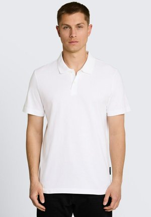 Młody mężczyzna ubrany w gładką białą koszulkę polo z krótkim rękawem i czarne spodnie, stojący na tle jasnoszarego tła.