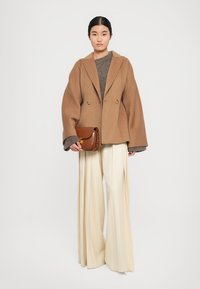Un manteau en laine marron croisé sur un pull gris, associé à un pantalon large crème, tenant un petit sac à bandoulière en cuir marron.