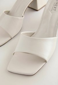Mules blanches à bout ouvert avec une large bride avant et un talon bloc bas sur fond clair.