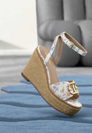 Kiilakorkosandaali, jossa on punottu korokepohja, kukkakuviolliset hihnat, kultainen solki ja nilkkahihna, asetettuna siniselle matolle harmaan tuolin eteen.