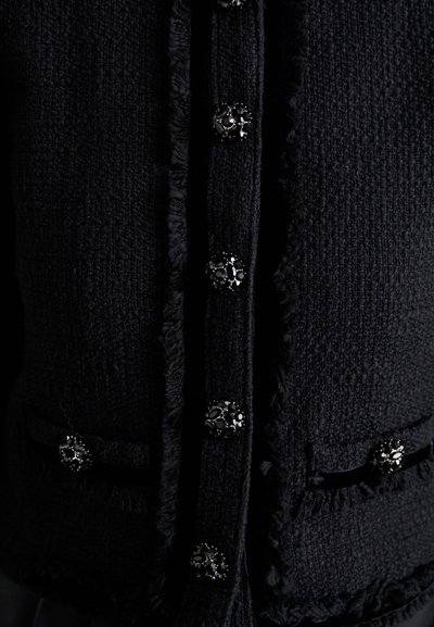 Zwarte tweed jas met een gestructureerd oppervlak, voorzien van rafelige randen en decoratieve knopen met zwarte en zilveren accenten. Twee voorzakken.
