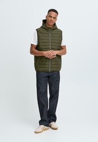 Grønn polstret vest med hette, med vertikale kviltede linjer, glidelås foran og lommer. Kombinert med mørke jeans og lyse joggesko.