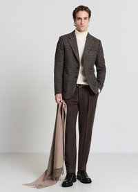 Braun kariertes Sakko über einem cremefarbenen Rollkragenpullover, kombiniert mit braunen Hosen. Hält einen beigen Schal, trägt schwarze glänzende Schuhe. Minimalistischer Hintergrund.