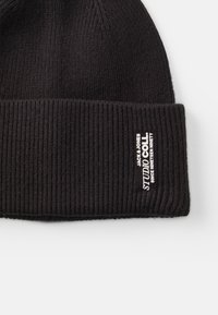 Zwarte gebreide beanie met een geribbelde textuur en een omgeslagen boord. Bevat een wit logo label op de boord. Eenvoudig, functioneel ontwerp.