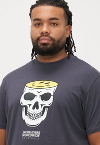 Σκούρο γκρι βαμβακερό μπλουζάκι με έντονο λευκό γραφικό κρανίο, επάνω στο οποίο υπάρχει κίτρινο χαμογελαστό προσωπάκι και το κείμενο "JACK & JONES WORLDWIDE ORIGINALS" από κάτω.
