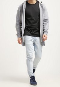 Felpa grigia con zip sopra una maglietta nera, jeans azzurri strappati e sneakers blu navy. Design semplice, look casual.