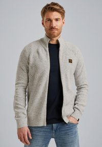 Muškarac s svijetlosmešom kosom i bradom, obučen u svijetlosivu zip-up duksericu preko tamnoplave majice i plavih traperica, stoji naslonjen na običnu sivu pozadinu.