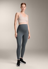 Svetlo siva športna modrček in temno siva visoko pasu legging, izdelana iz gladkega, raztegljivega materiala, v kombinaciji s črnimi športnimi čevlji.