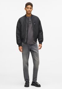 Juodas bombonešis su užtrauktuku priekyje, raištinėmis rankovėmis ir dviem krūtinės kišenėmis; derinamas su pilku marškinėliu, pilkomis džinsomis ir juodais batais.