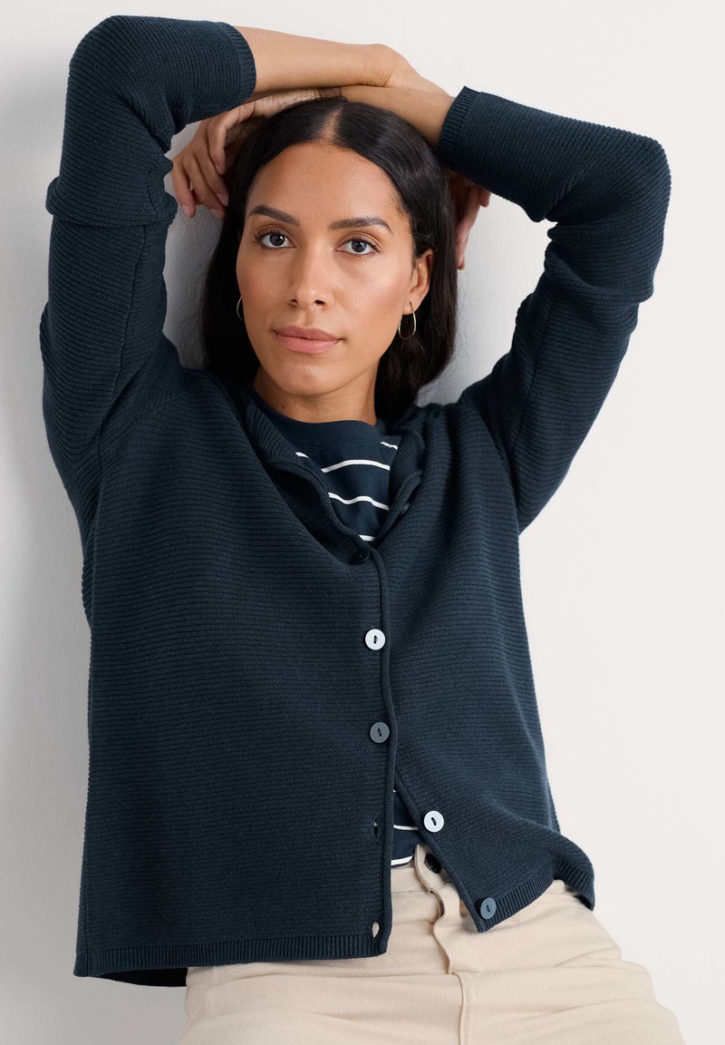Cardigan côtelé bleu marine avec encolure arrondie, fermeture par boutons et manches longues. Porté sur un haut rayé, il présente une texture subtile.