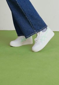 White sneakers with mint green accents, featuring a thick sole and perforated details, paired with dark denim flared pants on a green floor.