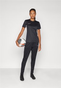 Svart Nike treningssett som inkluderer en t-skjorte med korte ermer og hvite detaljer, samt matchende bukser. Modellen holder en svart og hvit fotball.