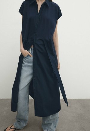 Femme portant une robe bleu marine sans manches boutonnée, ouverte, sur un jean bleu clair et des sandales noires, debout contre un mur blanc.