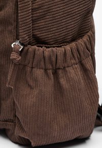 Sac à dos en velours côtelé brun avec des nervures verticales, poches latérales froncées et quincaillerie en argent pour la fermeture et les ajustements. Détail de surface texturé.