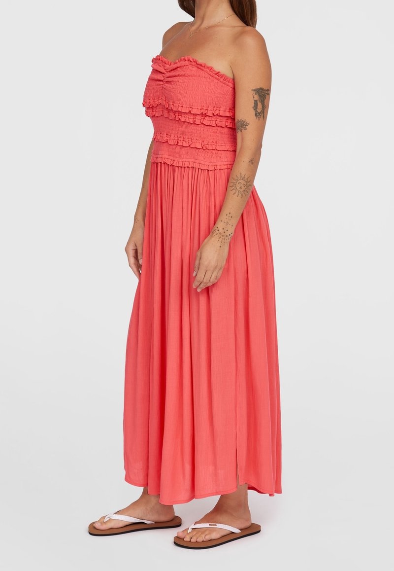 Robe maxi corail avec un corsage smocké et des accents volants, présentant une jupe fluide. Portée avec des tongs blanches, elle met en valeur une fente sur le côté.